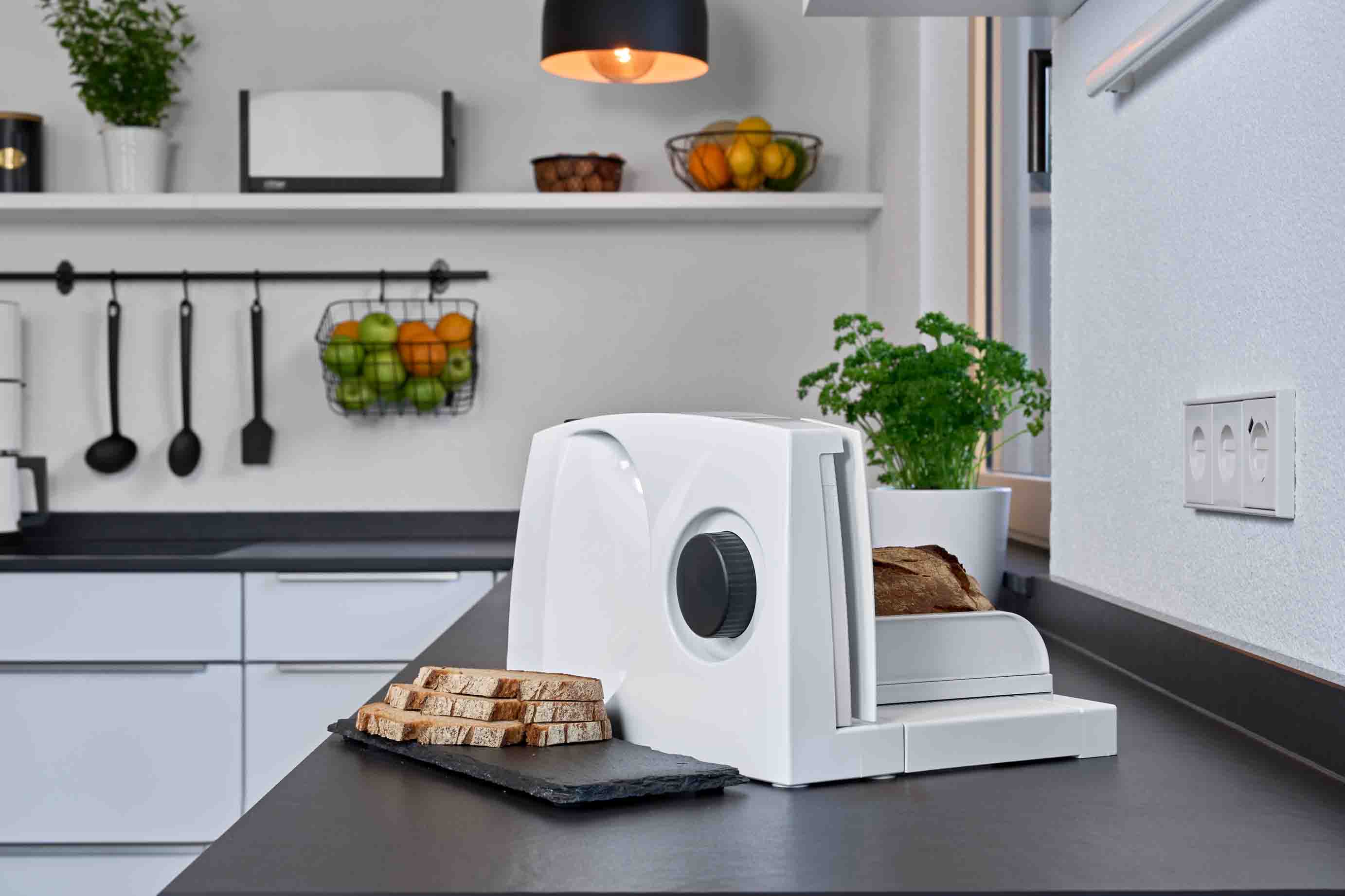 White food slicer on a dark kitchen worktop with chopped food and a pot of basil.