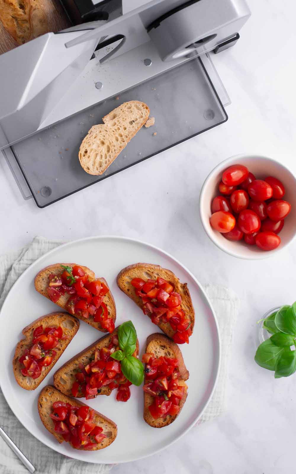 Frische Bruschetta mit Tomatenwürfeln und Basilikum auf gerösteten Brotscheiben, angerichtet auf einem weißen Teller vor einem ritter Allesschneider mit frisch geschnittenem Baguette