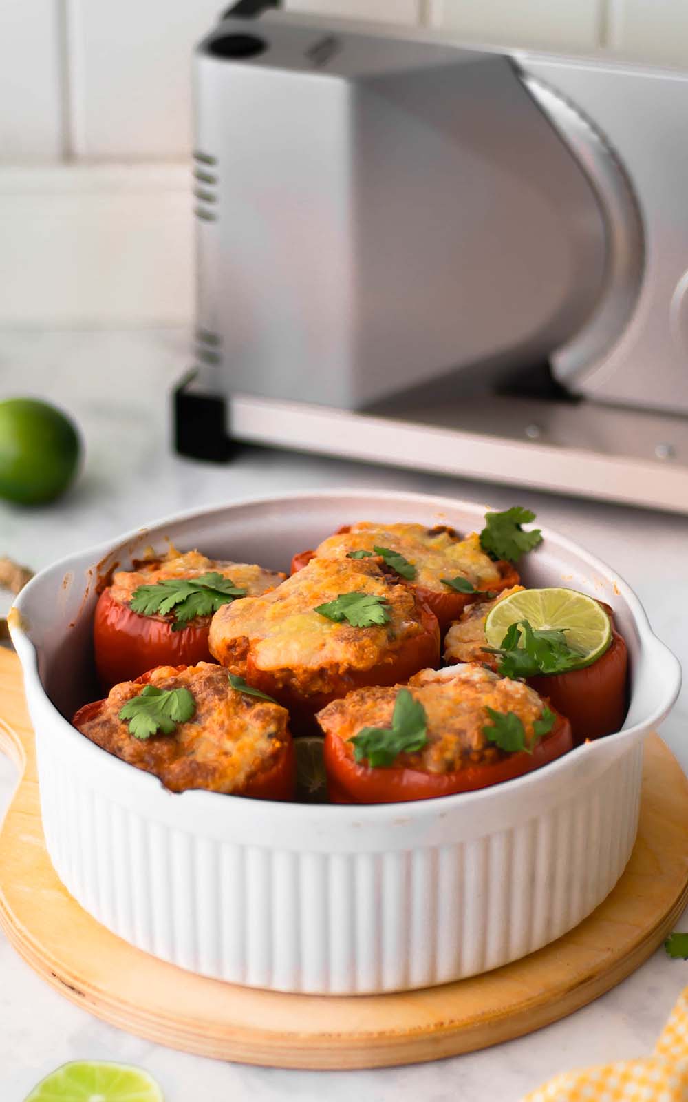 Auflaufform mit mehreren gefüllten Ofenpaprika, überbacken mit Käse und garniert mit frischem Koriander und Limettenscheiben. Im Hintergrund ein elektrischer Allesschneider