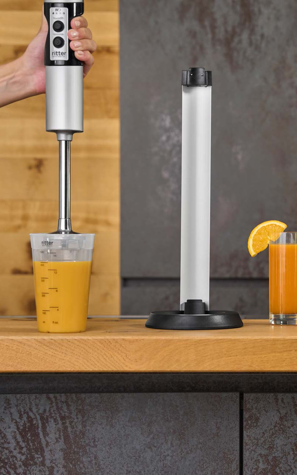Ein Bild von einem silbernen Stabmixer der einen Saft in einem Messbecher mixt und einem Glas gepressten Orangensaft, die in einer Küche stehen. Ein Bild von einem silbernen Stabmixer der einen Saft in einem Messbecher mixt und einem Glas gepressten Orangensaft, die in einer Küche stehen.