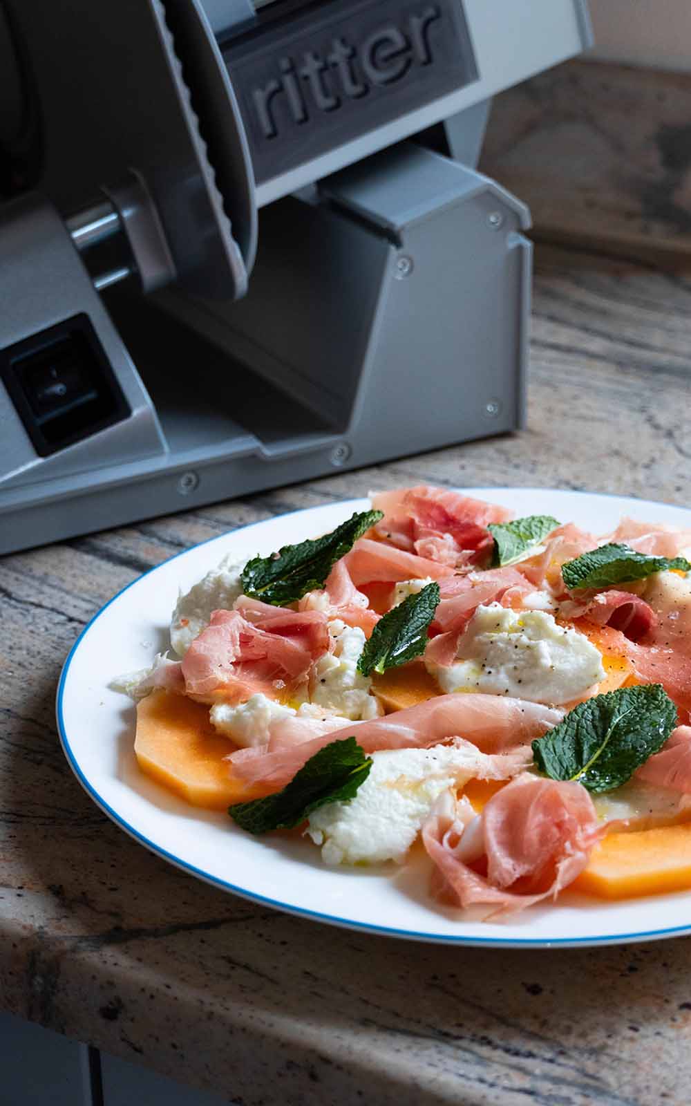 Melonensalat mit Prosciutto, Mozzarella und frischer Minze auf einem weißen Teller, daneben eine Aufschnittmaschine. Melonensalat mit Prosciutto, Mozzarella und frischer Minze auf einem weißen Teller, daneben eine Aufschnittmaschine.