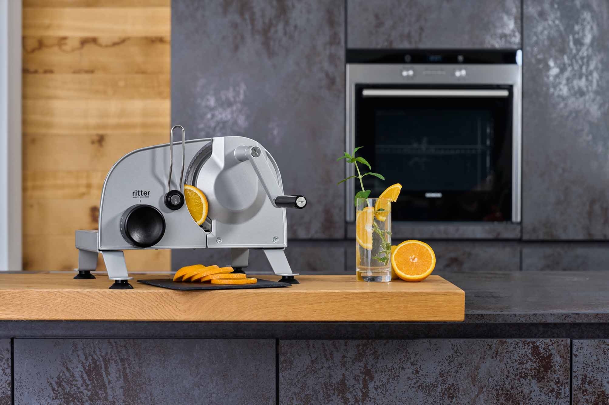 Silver manual food slicer on wooden worktop with sliced oranges and drink in modern kitchen
