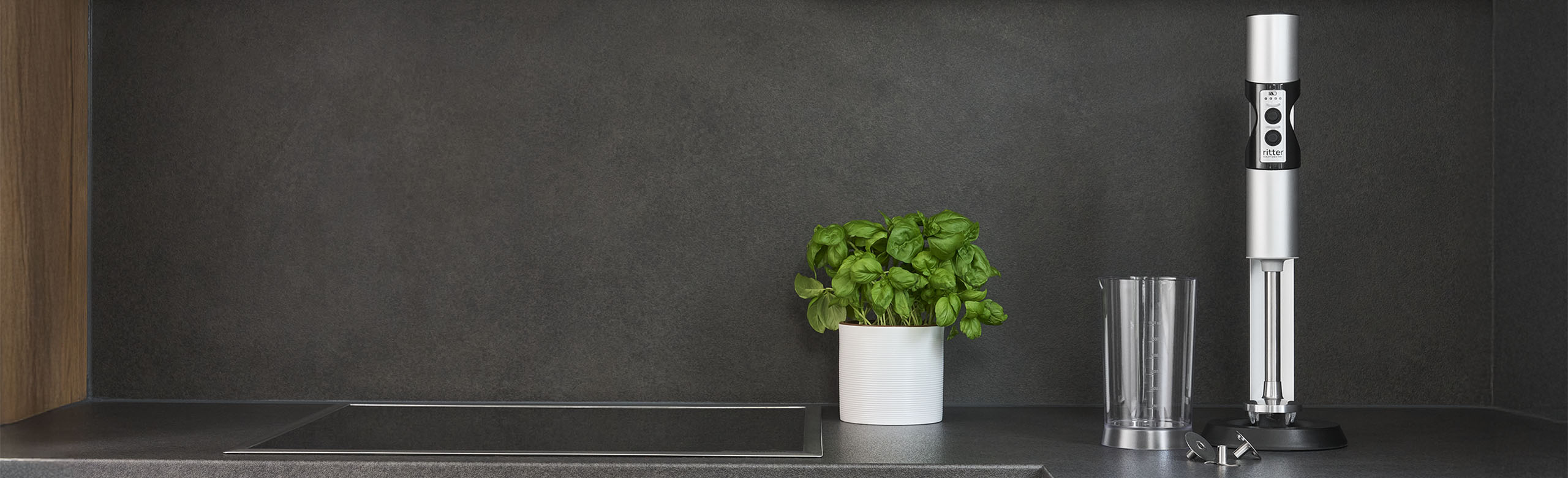 A picture of a silver hand blender, a filled measuring jug and a glass filled with juice in a kitchen.