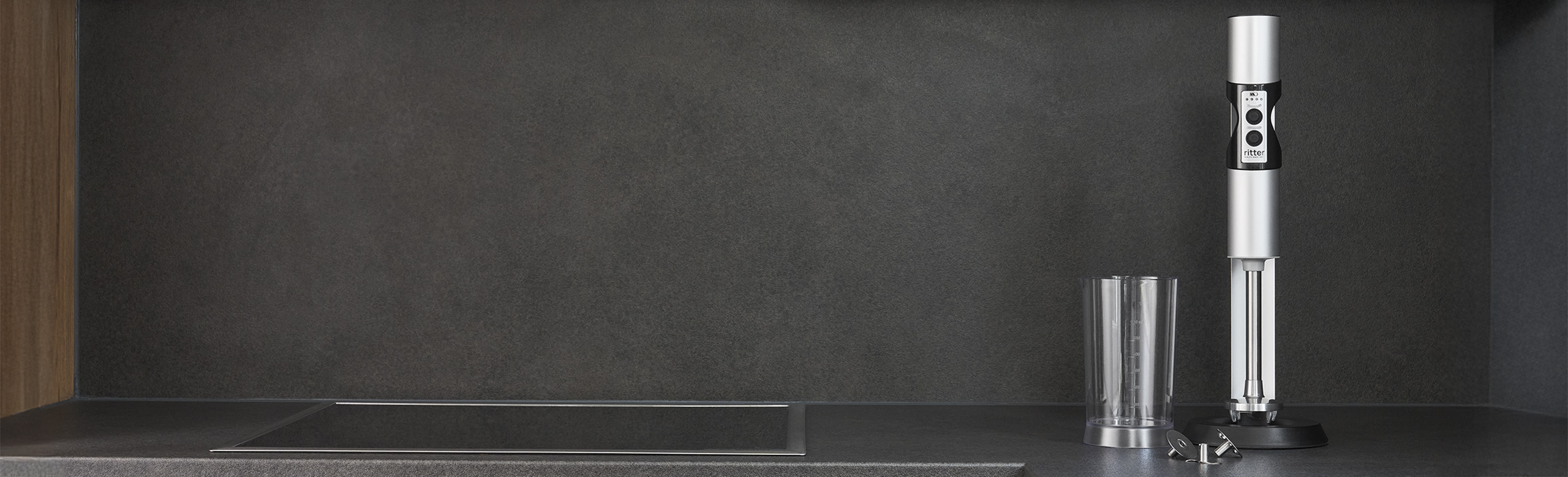 Ein Bild von einem silbernen Stabmixer, einem gefüllten Messbecher und einem, mit Saft gefüllten, Glas, in einer Küche.