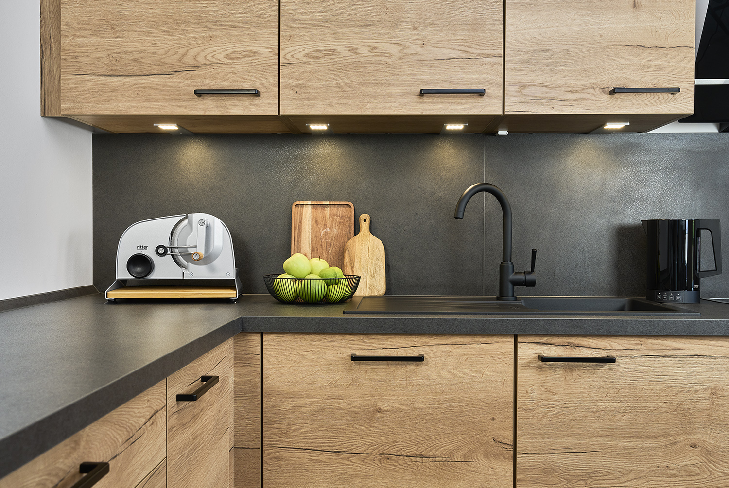Silver manual food slicer of the brand ritter with black knob, handle and bamboo board, on worktop with a bowl of apples to the right in modern kitchen