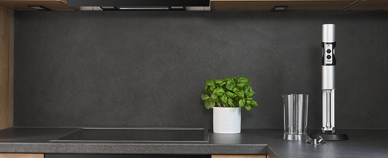 A picture of a silver hand blender, a filled measuring jug and a glass filled with juice in a kitchen.