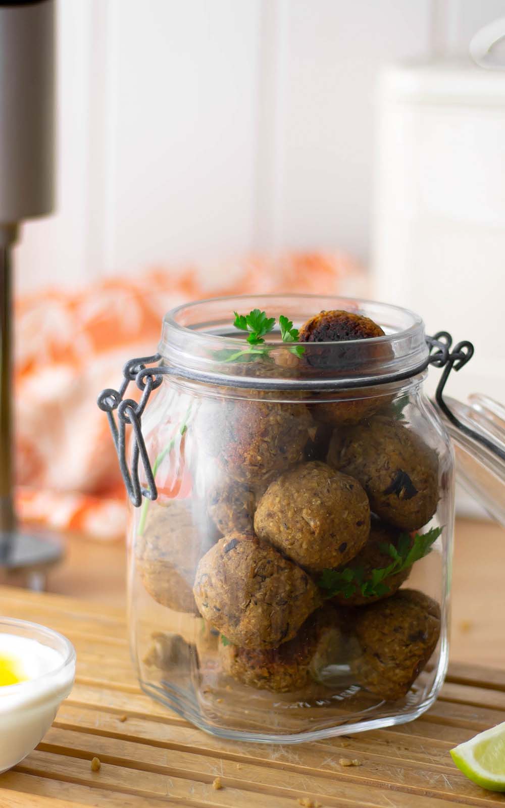 Ein Glas mit selbstgemachten veganen Falafel-Bällchen, garniert mit frischer Petersilie. Im Vordergrund steht eine kleine Schale mit Dip, daneben ein Limettenstück. Im Hintergrund ein Stabmixer und ein orange-weißes Küchentuch auf einer Holzoberfläche