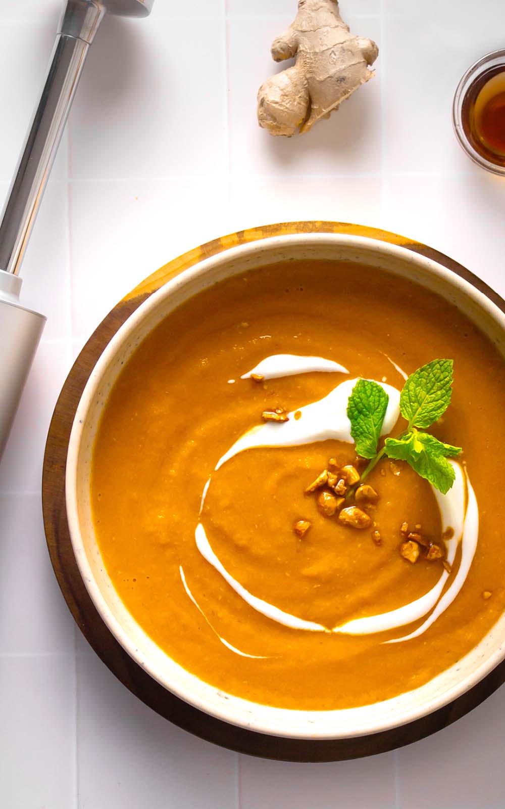 Schüssel mit cremiger roter Linsensuppe, garniert mit einem Klecks Joghurt, frischer Minze und gehackten Nüssen, daneben ein Stück Ingwer und ein Stabmixer auf weißem Untergrund. Schüssel mit cremiger roter Linsensuppe, garniert mit einem Klecks Joghurt, frischer Minze und gehackten Nüssen, daneben ein Stück Ingwer und ein Stabmixer auf weißem Untergrund.